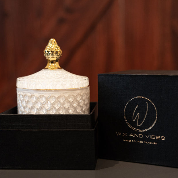  A luxurious white-patterned Wix and Vibes candle jar with a golden crown lid, placed beside its elegant black packaging box with the brand's logo in gold. sexy candles. masculine candles. black owned candle company. 